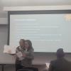 People giving a presentation, standing before and audience and in front of a screen. 