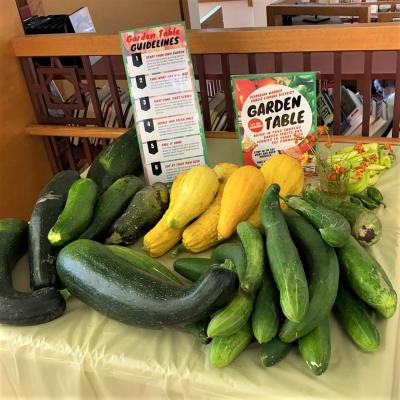 A variety of zucchini, yellow squash, cucumbers, and a small vase of orange flowers sits on a yellow tablecloth with Garden Table instructions. 