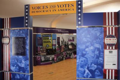 Image of an arch entryway into the Voices and Votes: Democracy in America exhibition. The image features the title of the exhibition and a glimpse of the voting rights display panel. 