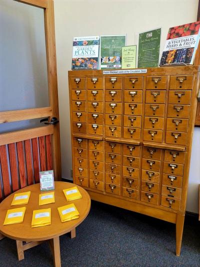 A photo of a free-standing card catalog and small table. The card catalog is filled with seed packets for library card holders to use in their home gardens. 