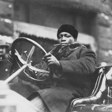 Black man sitting behind a wheel of an old convertible car with a cigar in his mouth