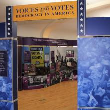 Image of an arch entryway into the Voices and Votes: Democracy in America exhibition. The image features the title of the exhibition and a glimpse of the voting rights display panel. 